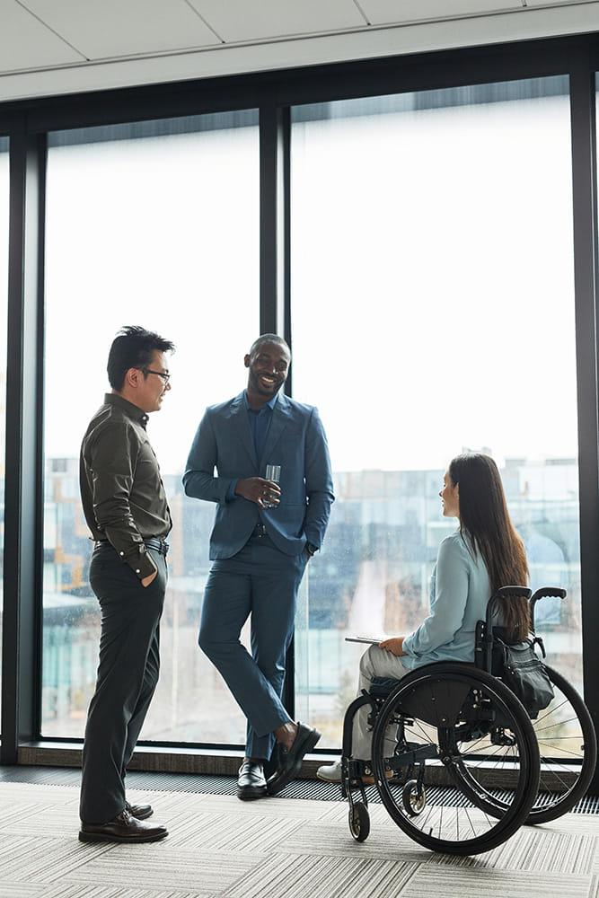 A group of three diverse professionals engaged in a conversation near large floor-to-ceiling windows in a modern office setting. The group includes a man in a suit leaning casually against the window with a glass in hand, another man wearing glasses and business attire, and a woman in a wheelchair holding a tablet. The scene reflects a professional and inclusive environment, with natural light streaming in from the windows.