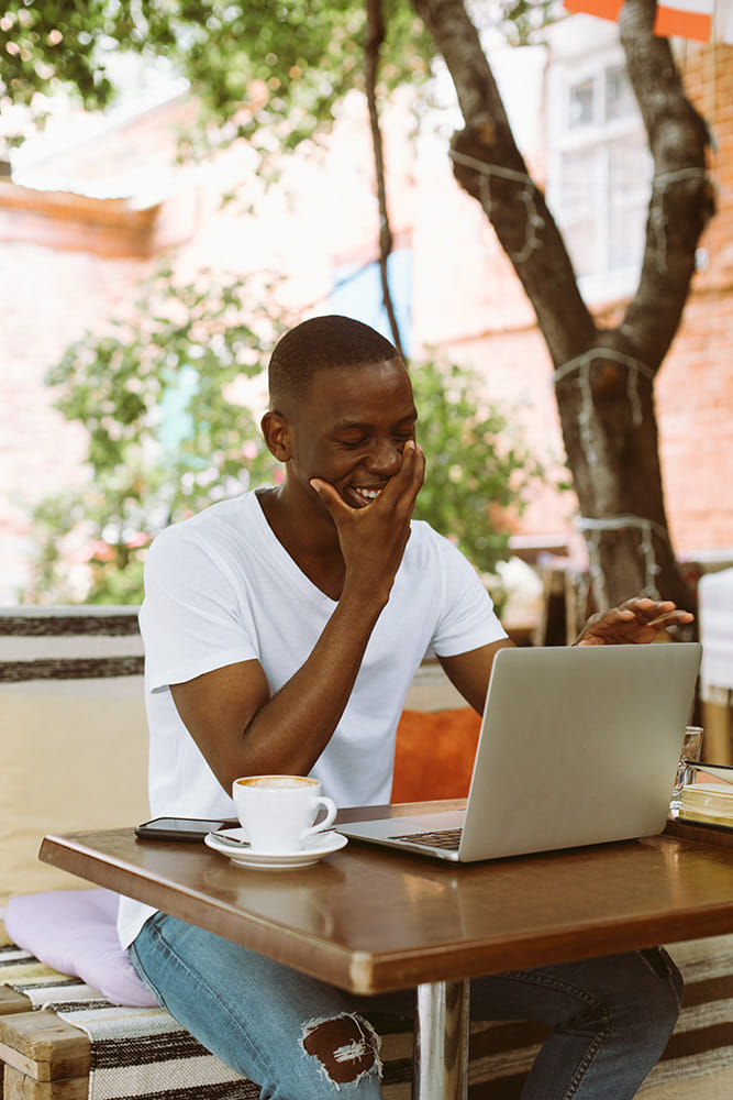 A young man wearing a white t-shirt and ripped jeans, sits at an outdoor café table with a laptop in front of him. He is smiling and holding his hand to his face, looking engaged with what is on the screen. A cup of coffee is placed next to the laptop, and the setting is cozy and surrounded by greenery, with soft sunlight filtering through the trees.